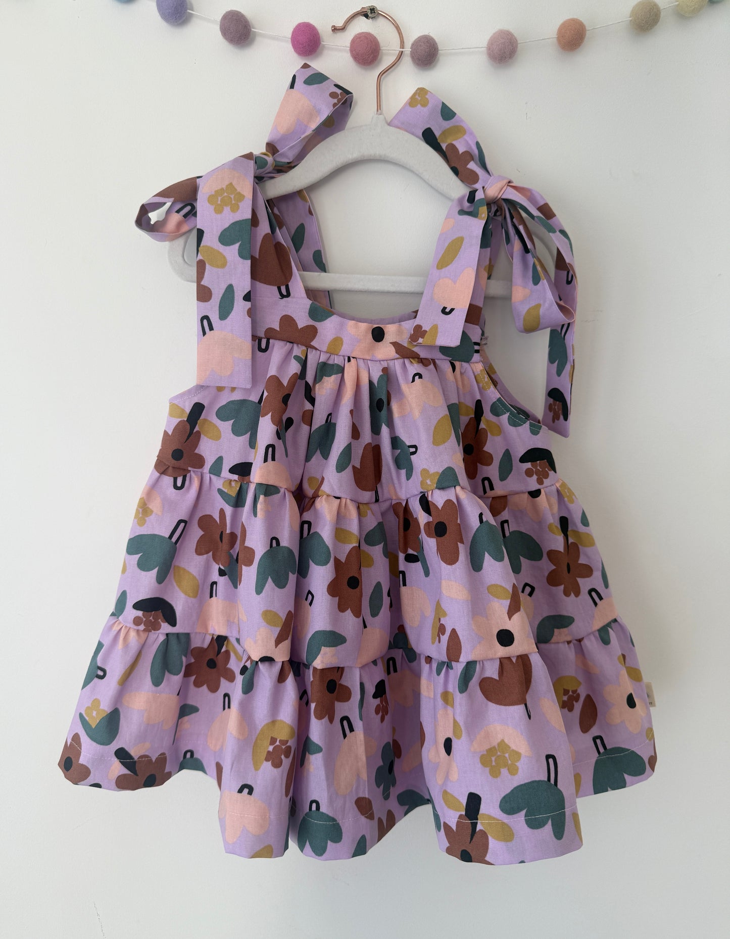 A toddler girl's tiered 100% cotton ruffle dress in lavender purple with a whimsical floral and abstract print, hanging on a wooden hanger against a white wall.