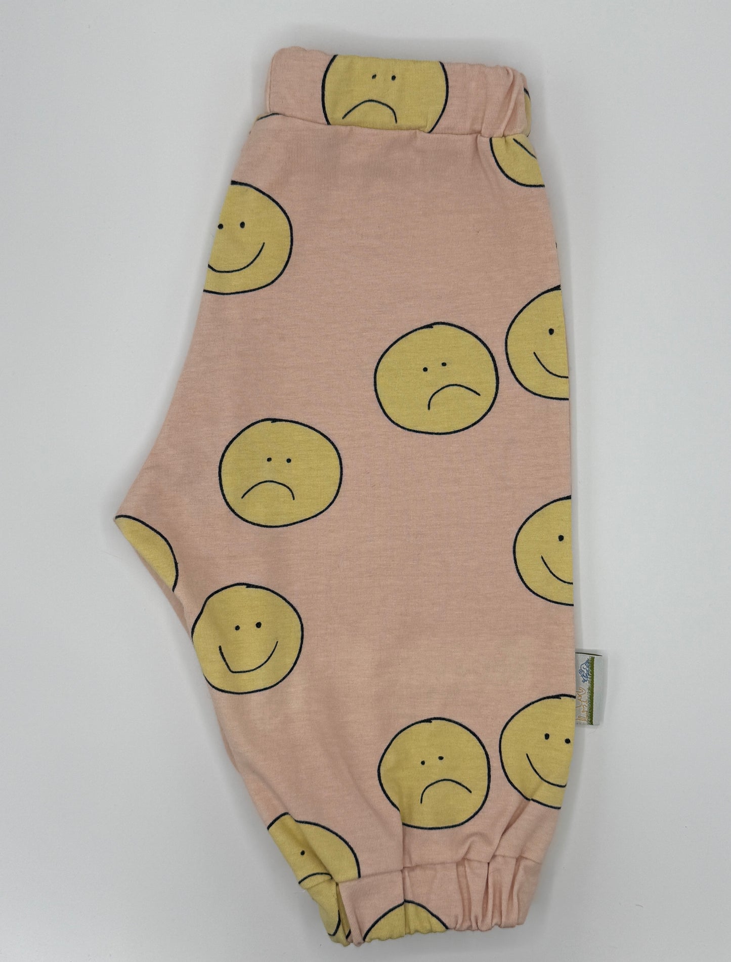 Pink unisex sweatpants with happy/sad faces in yellow with black outline. They have elasticated waist and cuffs 