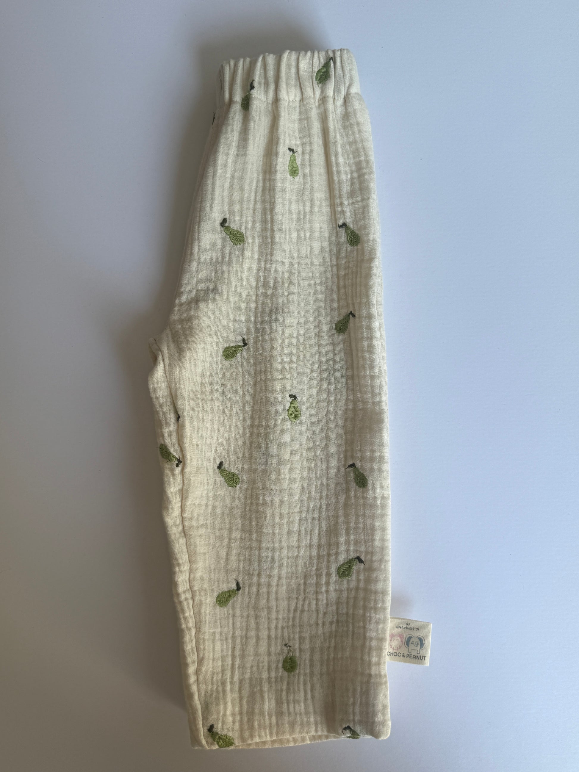 A pair of cream-colored, textured gauze baby pants with a green pear print, laid flat on a light gray background. A small brand tag is visible near the bottom right hem.